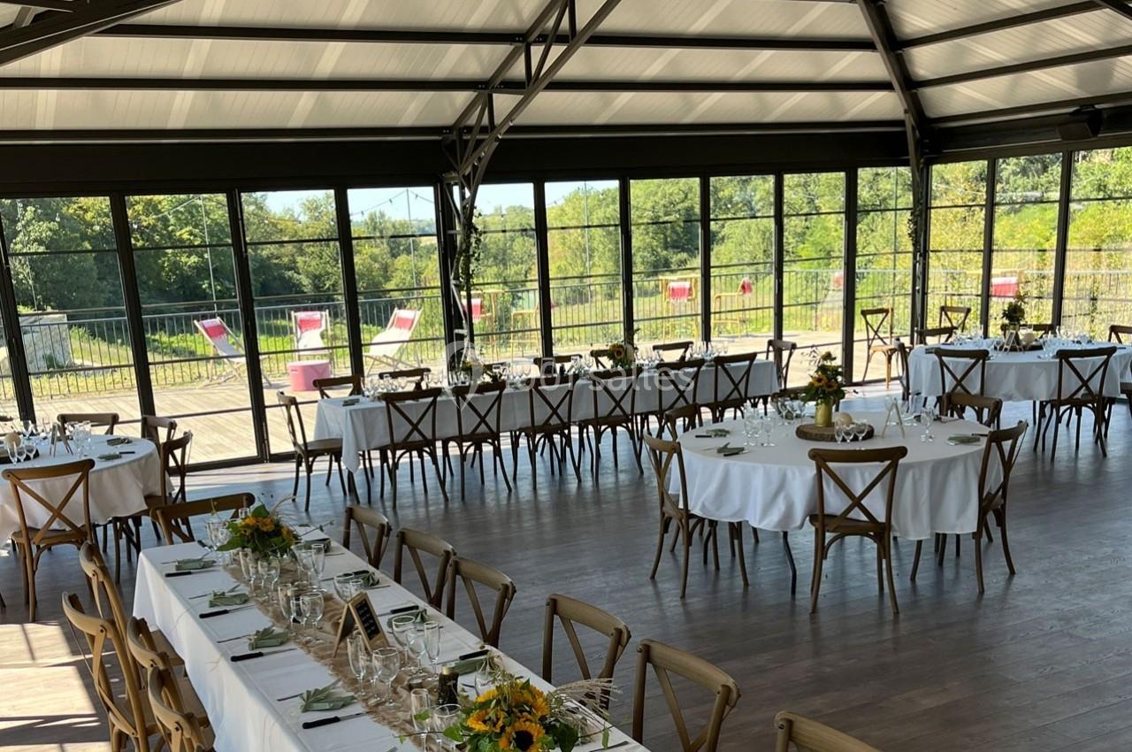 Salle lumineuse avec de grandes baies vitrées, tables dressées avec nappes blanches et bouquets de fleurs jaunes.