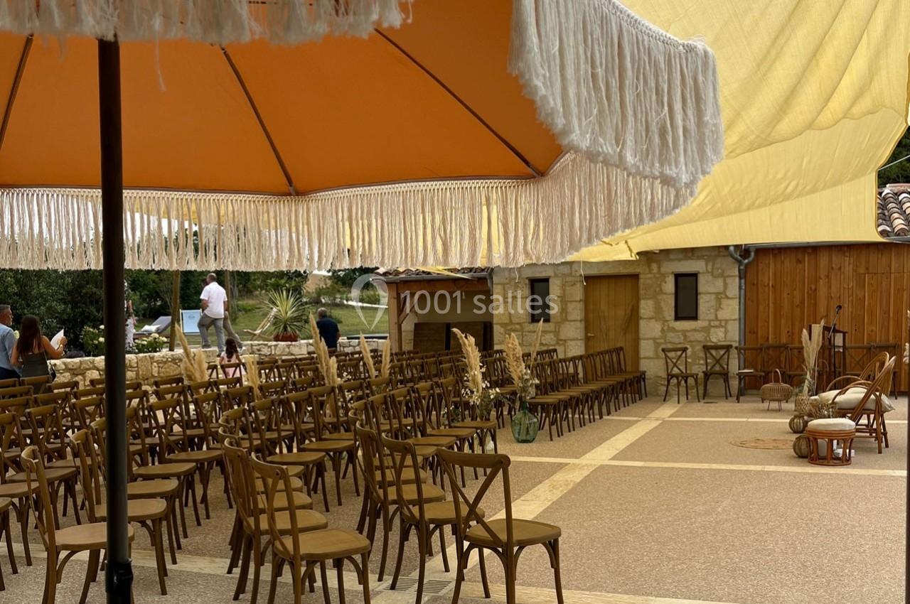 Chaises en bois disposées sous des parasols et une toile tendue dans une cour en pierre, préparée pour un événement.