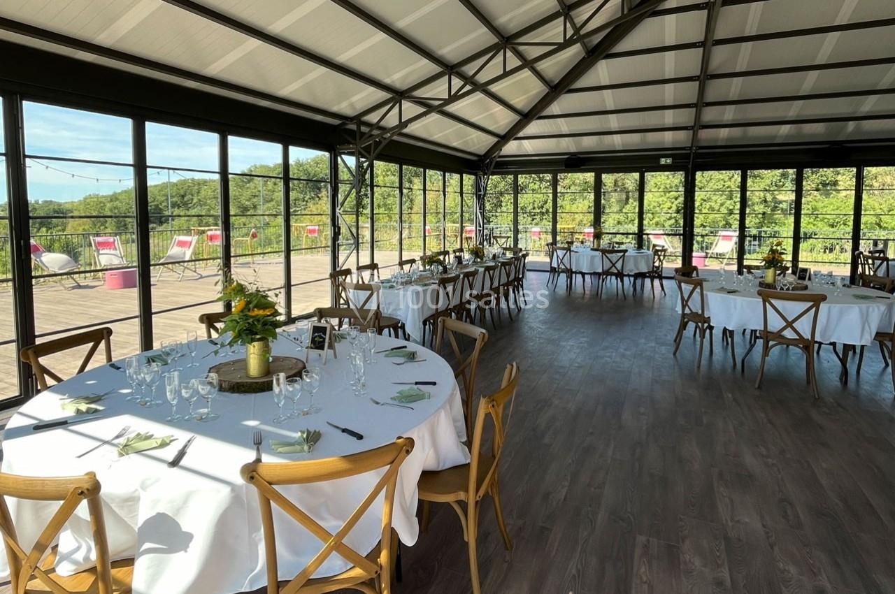 Salle lumineuse avec grandes baies vitrées, tables rondes dressées avec nappes blanches et vue sur un espace extérieur…