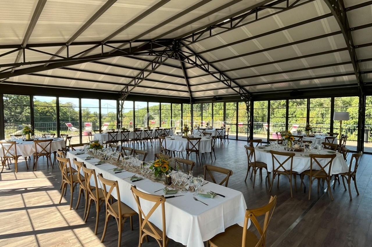 Salle lumineuse avec tables dressées pour un repas, grandes baies vitrées offrant une vue sur un paysage verdoyant.