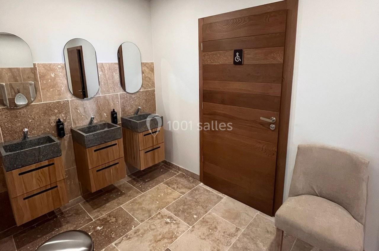 Salle de bain avec trois lavabos, miroirs ovales, sol en pierre et porte en bois avec pictogramme d'accessibilité.