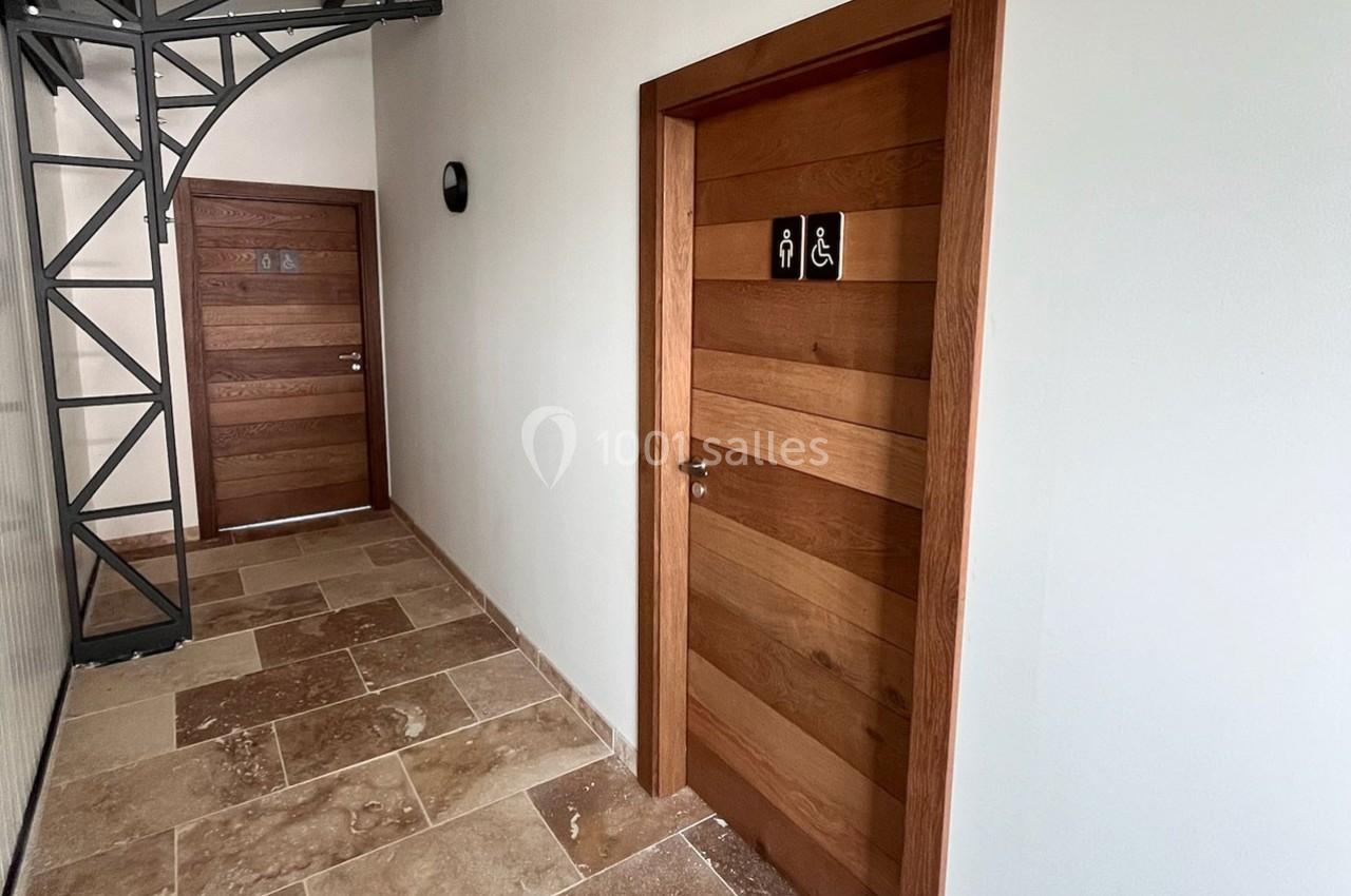 Porte en bois avec pictogrammes indiquant des toilettes accessibles, située dans un couloir au sol en pierre.