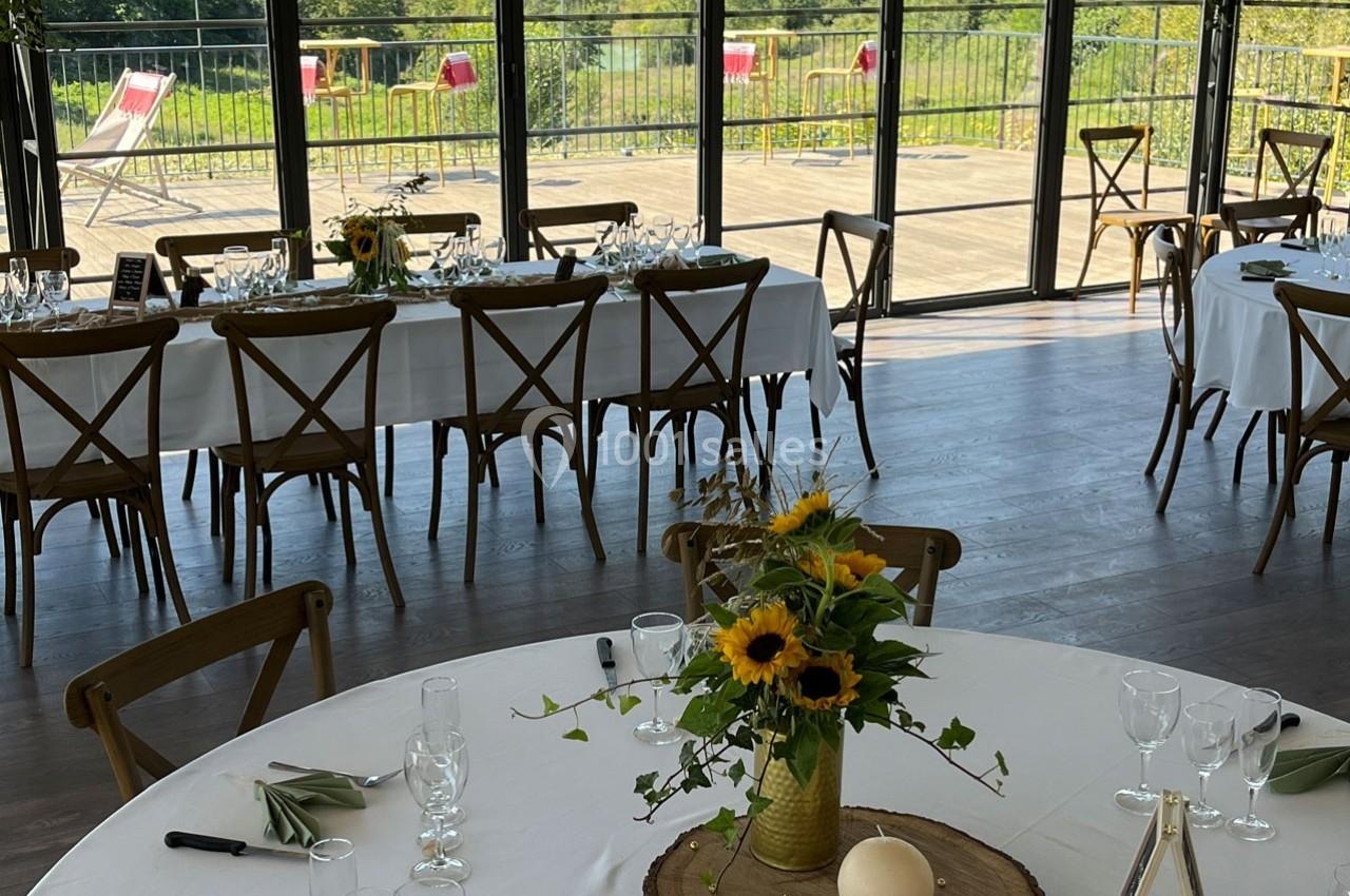 Salle lumineuse avec tables rondes et rectangulaires décorées de tournesols, donnant sur une terrasse et un paysage…