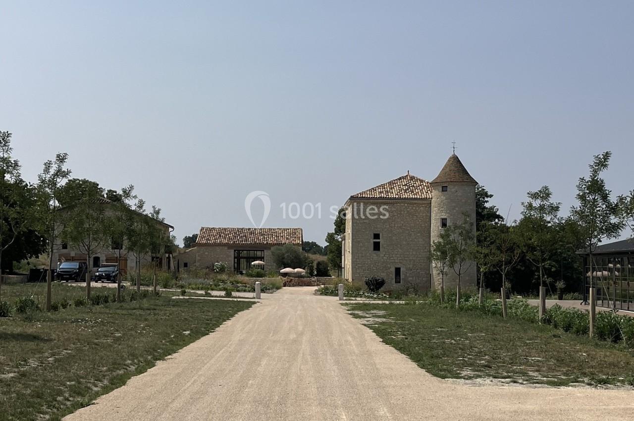 Allée bordée d'arbres menant à des bâtiments en pierre, dont une tour, sous un ciel dégagé.