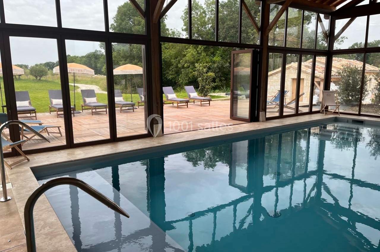 Piscine intérieure avec grandes baies vitrées donnant sur une terrasse en bois, des transats et un jardin verdoyant.