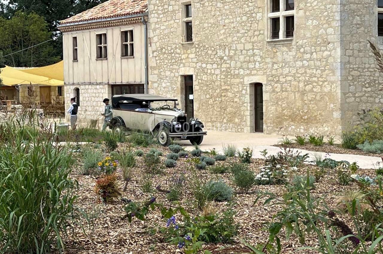 Voiture ancienne stationnée devant un bâtiment en pierre, entouré d'un jardin avec des plantes et des fleurs.
