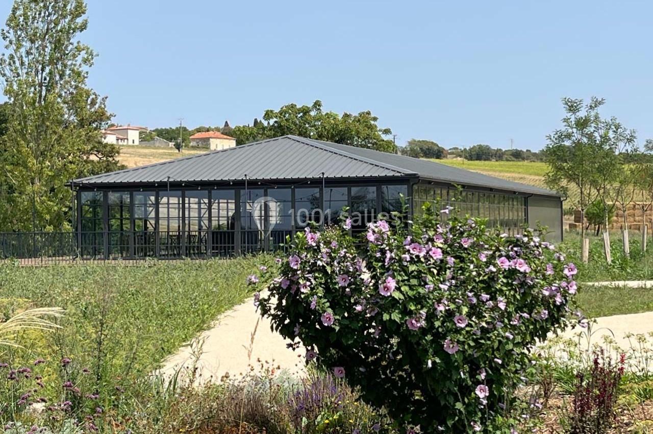 Vue d'un bâtiment moderne avec une structure en verre et métal, entouré de verdure et de fleurs violettes.