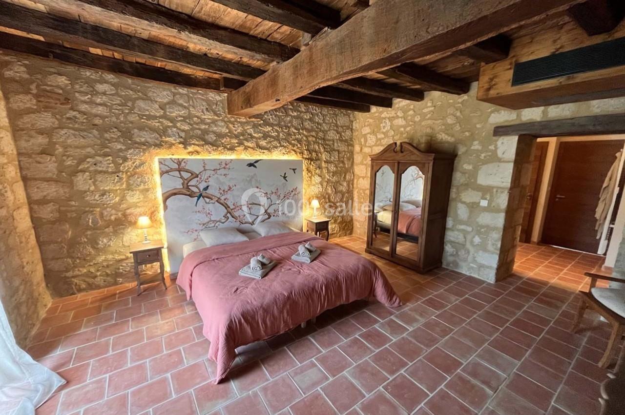 Chambre rustique avec murs en pierre, lit double, couvre-lit rose, armoire en bois et poutres apparentes au plafond.