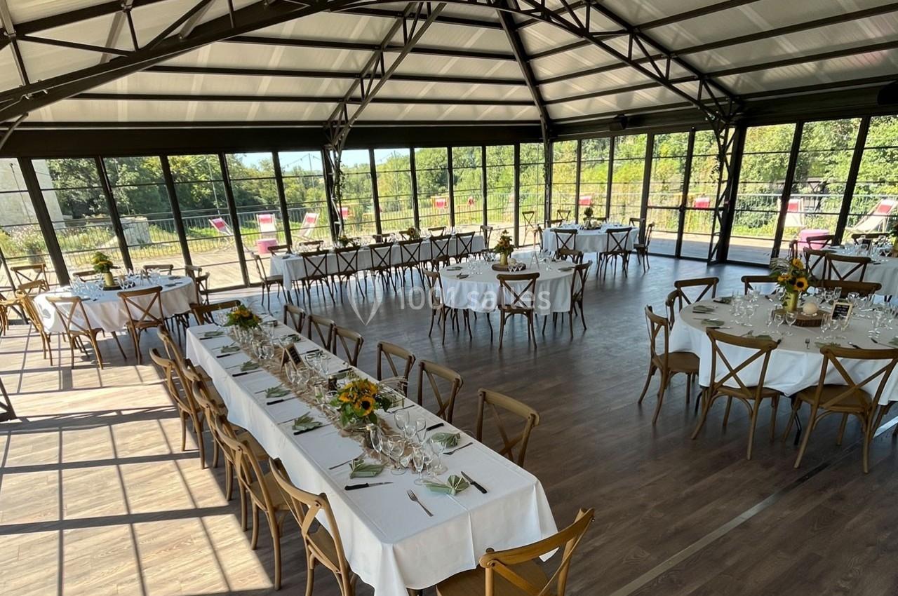 Salle lumineuse avec de grandes baies vitrées, tables dressées pour un repas, décorations florales sur les tables.