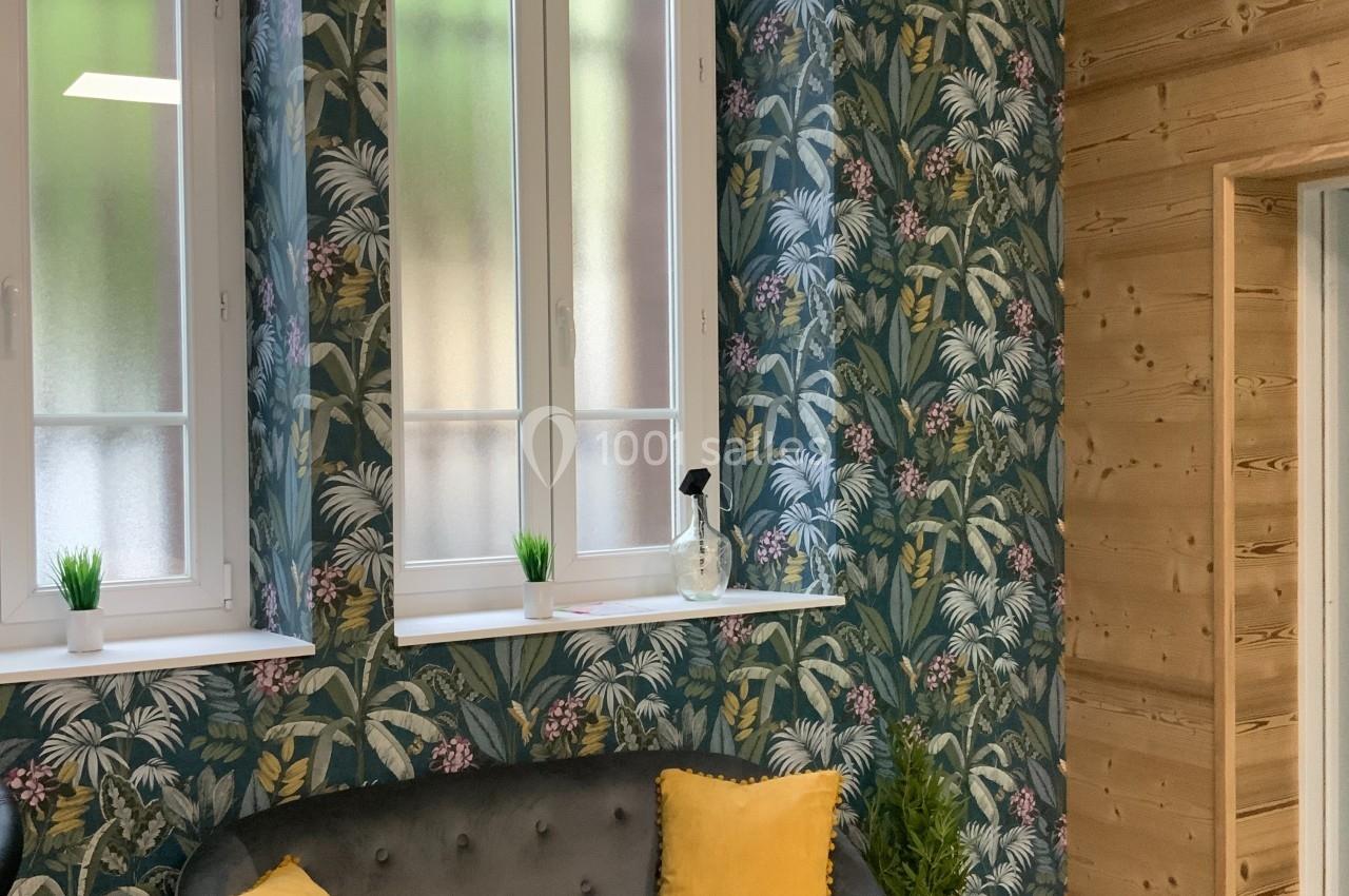Salon avec canapé gris, coussins jaunes, mur tapissé de motifs tropicaux, lambris en bois et fenêtres vitrées.