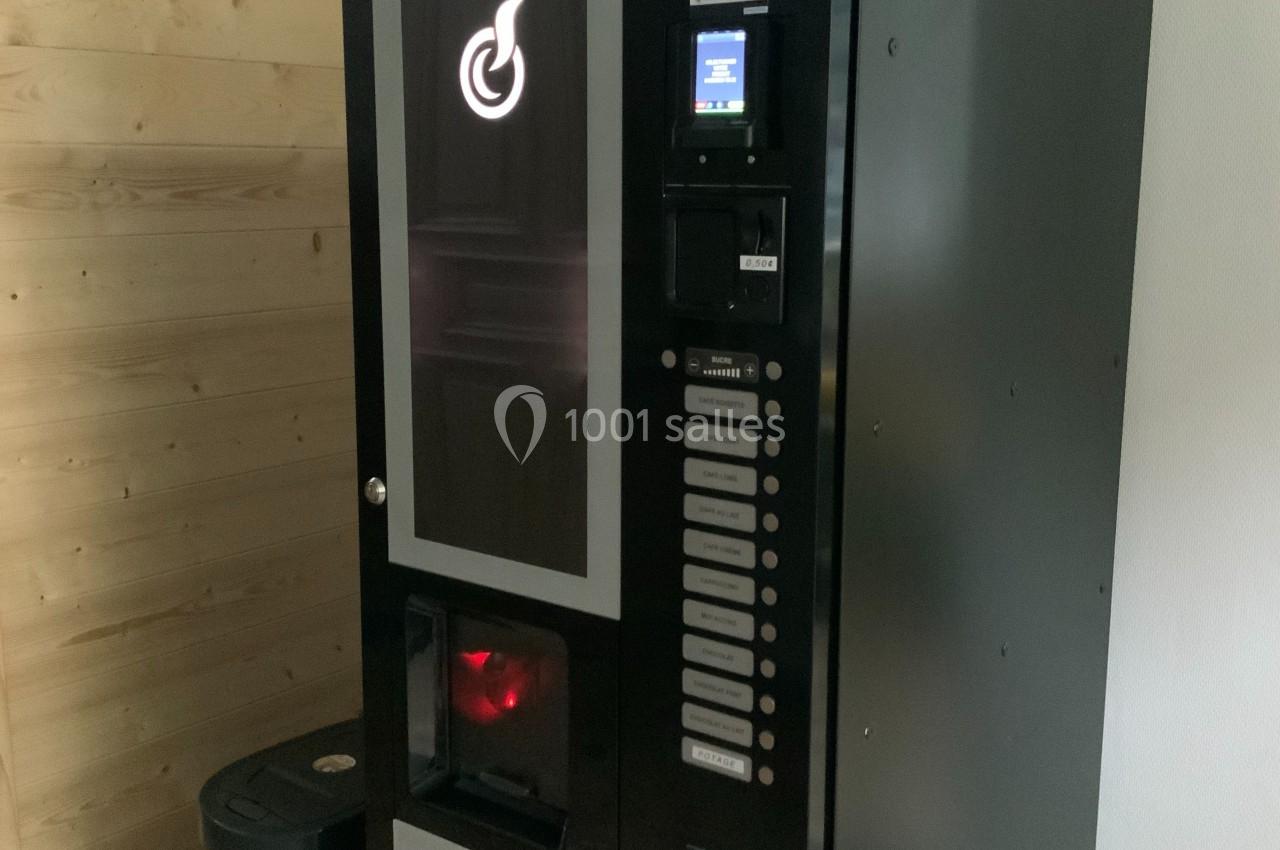 Distributeur automatique de boissons chaudes noir, installé près d'un mur en bois clair et d'une poubelle noire.