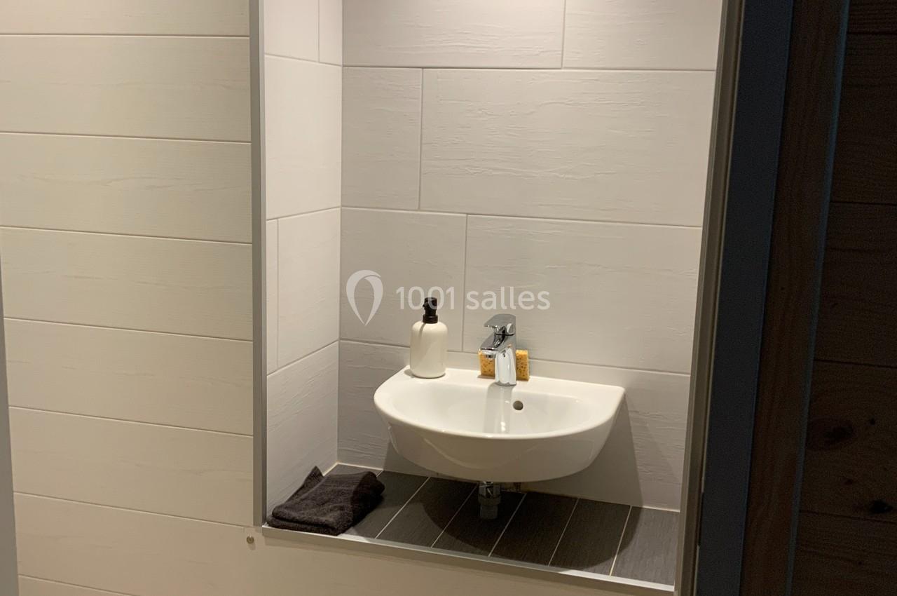 Lavabo blanc suspendu dans une alcôve carrelée, éclairée par un spot, avec accessoires et serviette posée à côté.