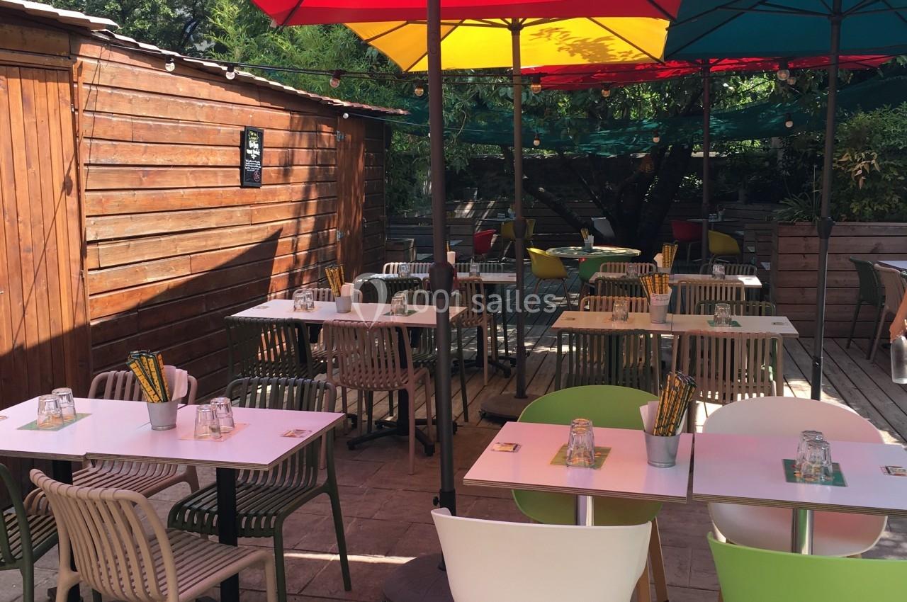 Terrasse ensoleillée avec tables dressées, chaises colorées et parasols rouges et verts, entourée de verdure et d'une…