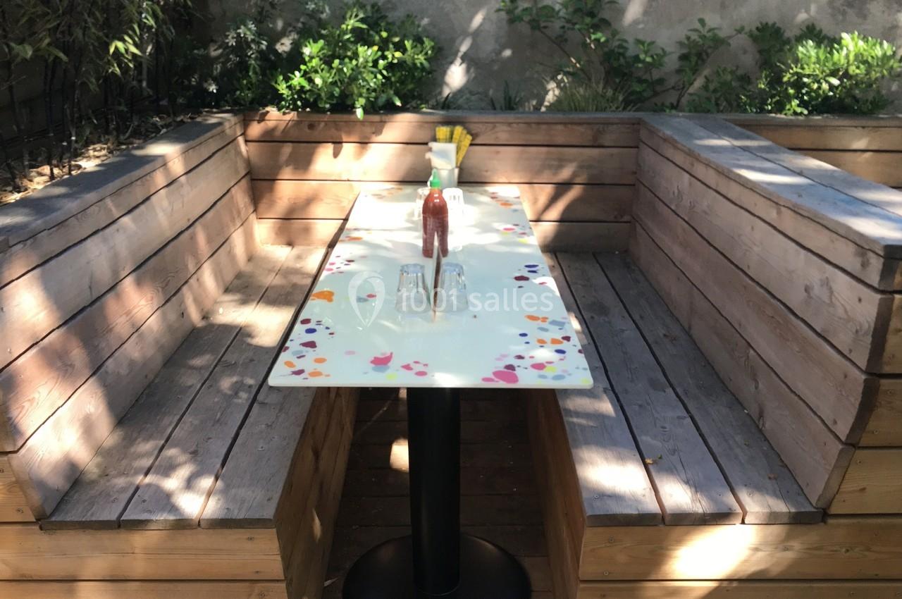 Banquette en bois entourant une table étroite avec une nappe blanche, située dans un jardin ombragé.