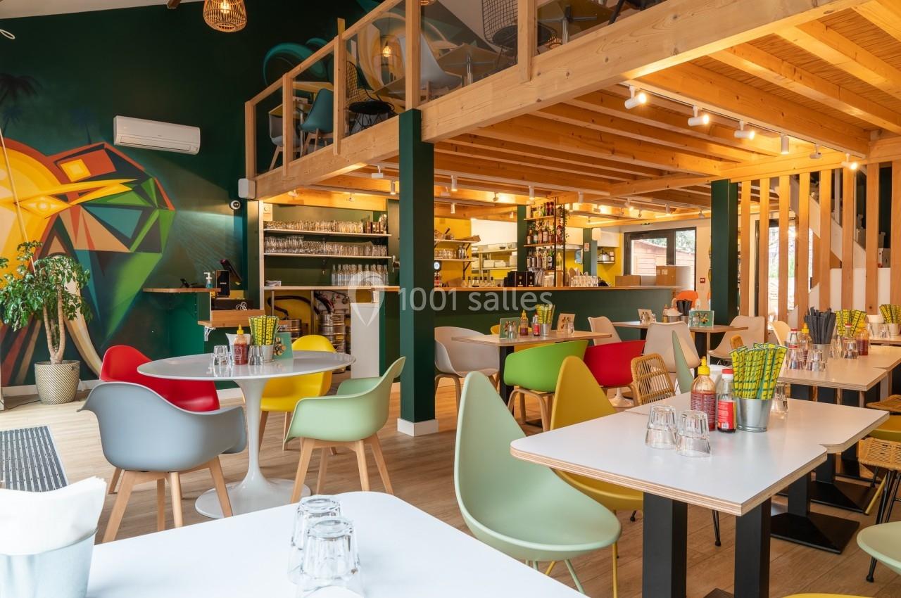 Salle de restaurant moderne avec mobilier coloré, mezzanine en bois, et mur décoré d'une fresque artistique.