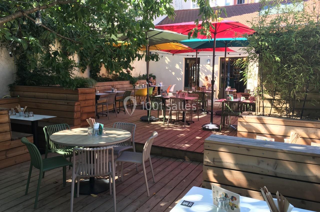 Terrasse en bois avec tables et chaises colorées, ombragée par des arbres et des parasols multicolores.