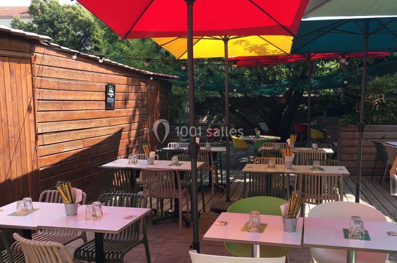 Terrasse extérieure avec tables et chaises colorées, ombragée par des parasols et entourée de verdure.