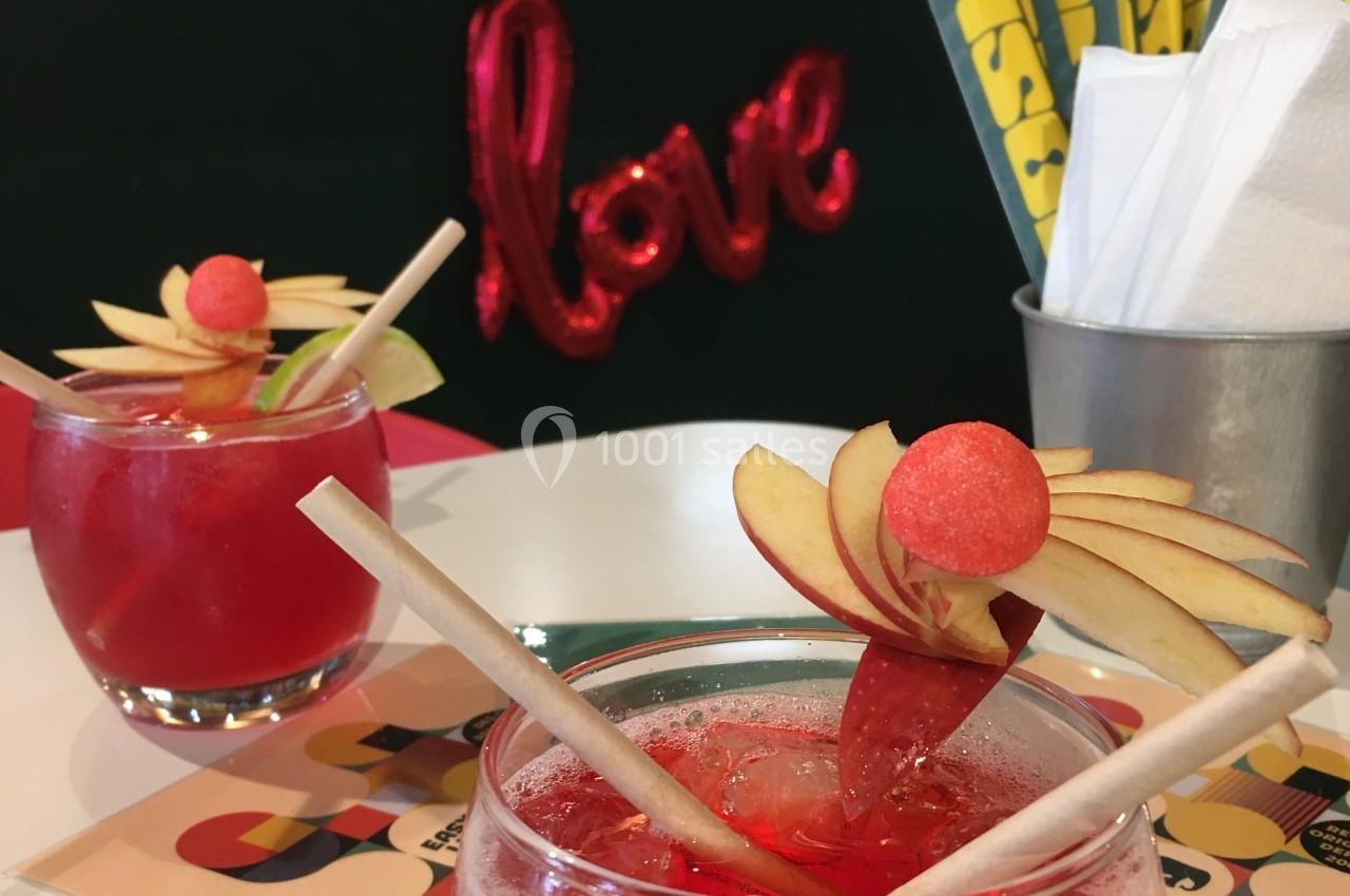Deux verres de cocktail rouge décorés de tranches de pomme et de bonbons, avec une décoration ’love’ en arrière-plan.
