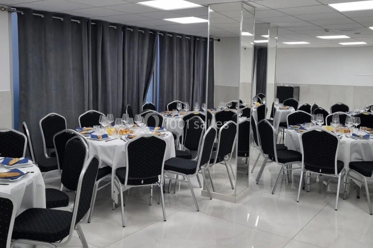 Salle de réception avec tables rondes dressées, nappes blanches, chaises noires et rideaux gris foncé.