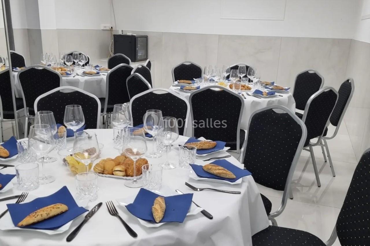 Tables rondes dressées avec nappes blanches, serviettes bleues, couverts, verres et viennoiseries dans une salle lumineuse.
