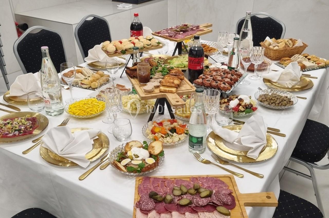 Table dressée avec divers plats, charcuteries, pains, salades, boissons et couverts, dans une salle lumineuse.