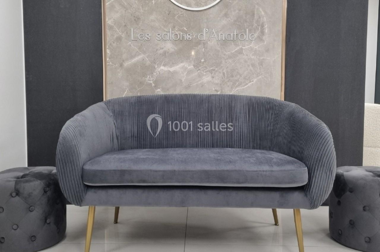 Canapé gris en velours côtelé avec pieds dorés, encadré par deux poufs capitonnés, devant un mur en marbre.