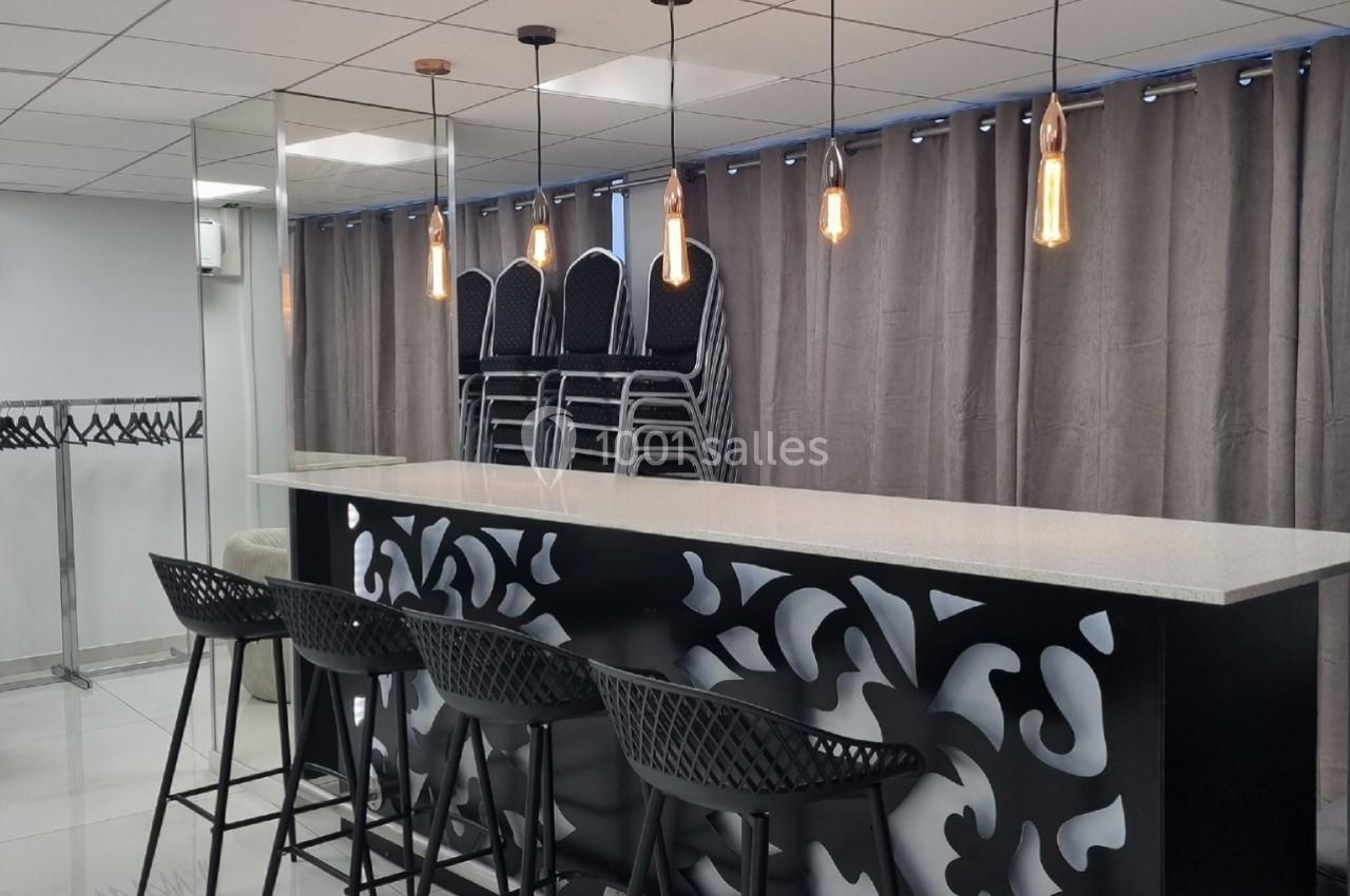 Comptoir moderne avec tabourets noirs, suspensions lumineuses et chaises empilées devant un rideau gris.