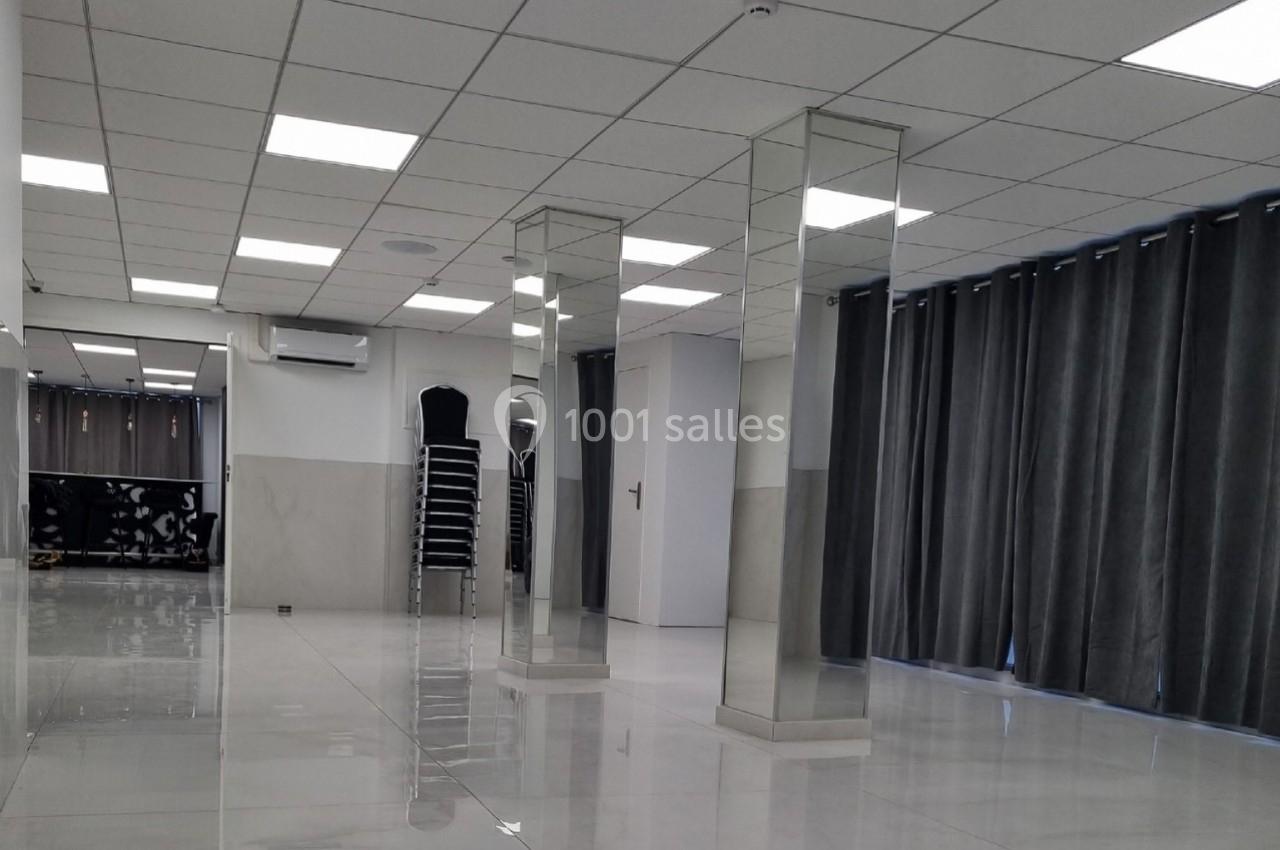 Salle vide avec sol brillant, murs clairs, colonnes en miroir, rideaux gris et chaises empilées dans un coin.