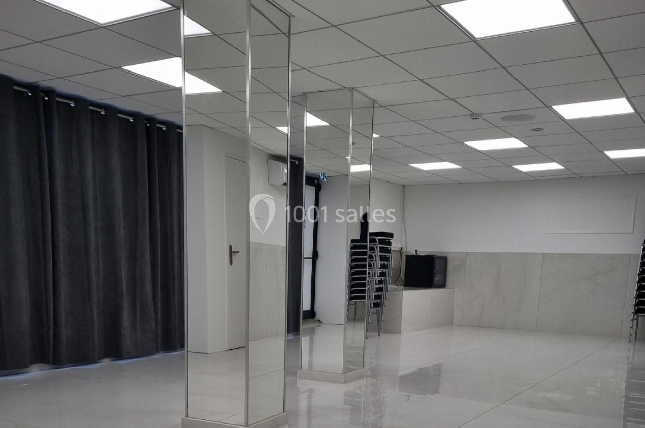 Salle lumineuse avec sol en carrelage brillant, colonnes recouvertes de miroirs, rideaux gris et chaises empilées au fond.