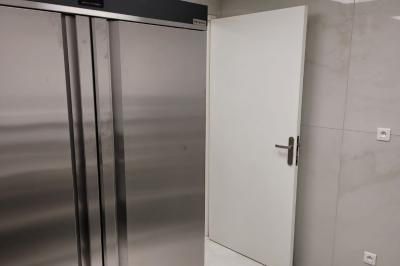 Réfrigérateur en acier inoxydable à double porte dans une pièce aux murs carrelés, avec une porte blanche ouverte.