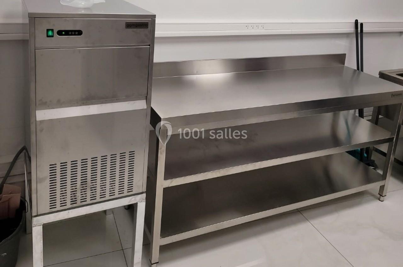 Machine à glaçons en acier inoxydable à côté d'une table de travail métallique dans une pièce au sol carrelé.