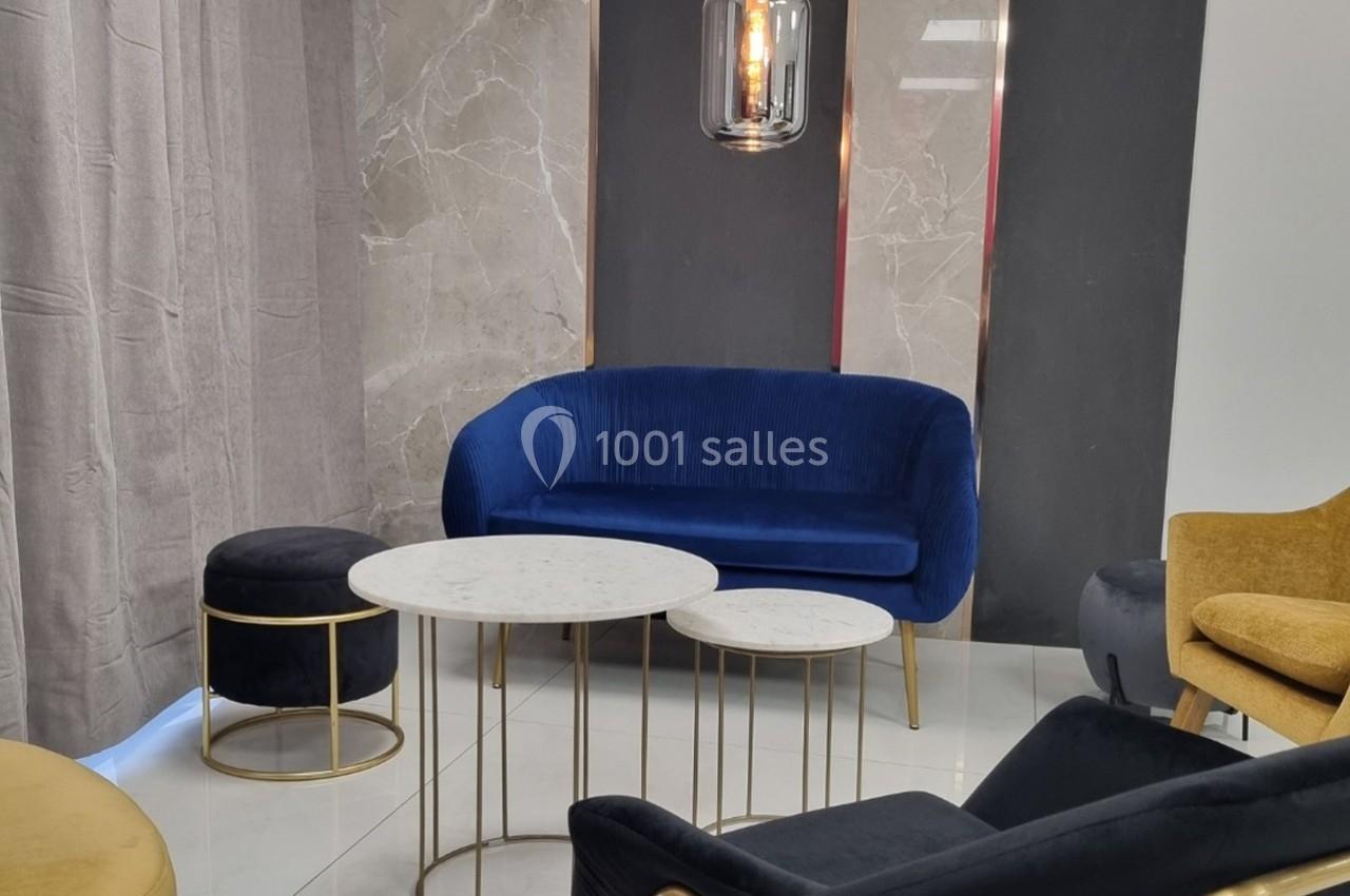 Salon moderne avec canapé bleu, fauteuils jaunes, tables basses en marbre et éléments dorés sur un sol blanc.