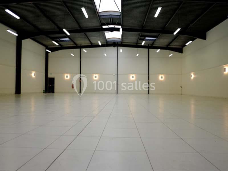 Intérieur d'un grand entrepôt vide avec sol carrelé blanc, murs clairs et plafond métallique éclairé par des néons.