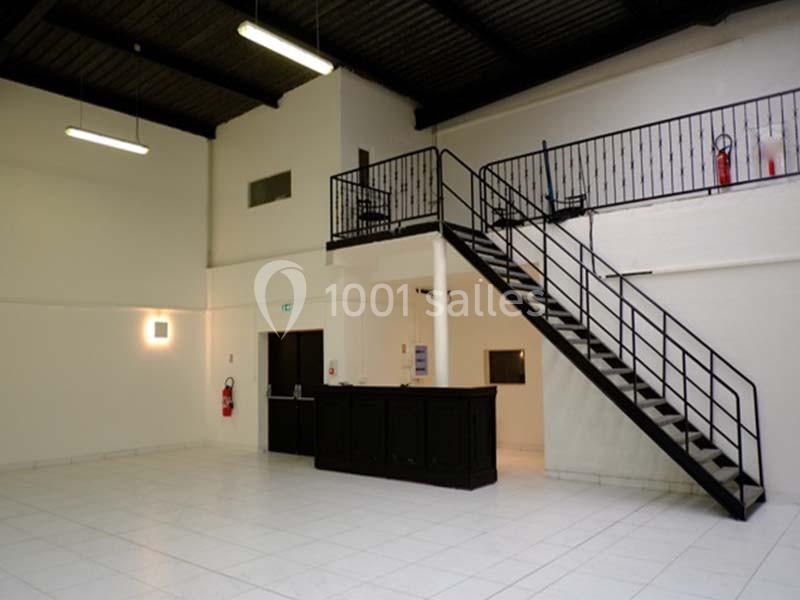 Intérieur d'un espace lumineux avec mezzanine, escalier métallique, comptoir noir et sol en carrelage blanc.