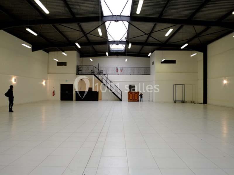 Grand espace intérieur avec sol carrelé blanc, plafond métallique, mezzanine et éclairage suspendu.