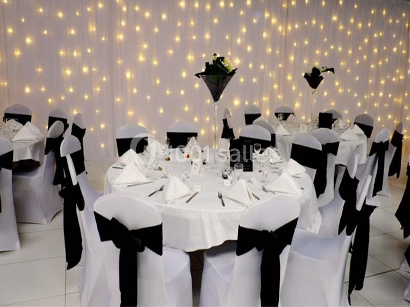 Salle décorée pour un événement, avec des tables rondes dressées, nappes blanches et chaises ornées de nœuds noirs.