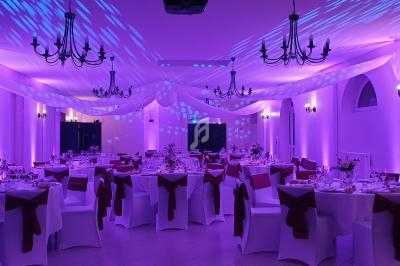 Salle de réception décorée avec des tables rondes, nappes blanches, chaises ornées de nœuds rouges et éclairage violet.