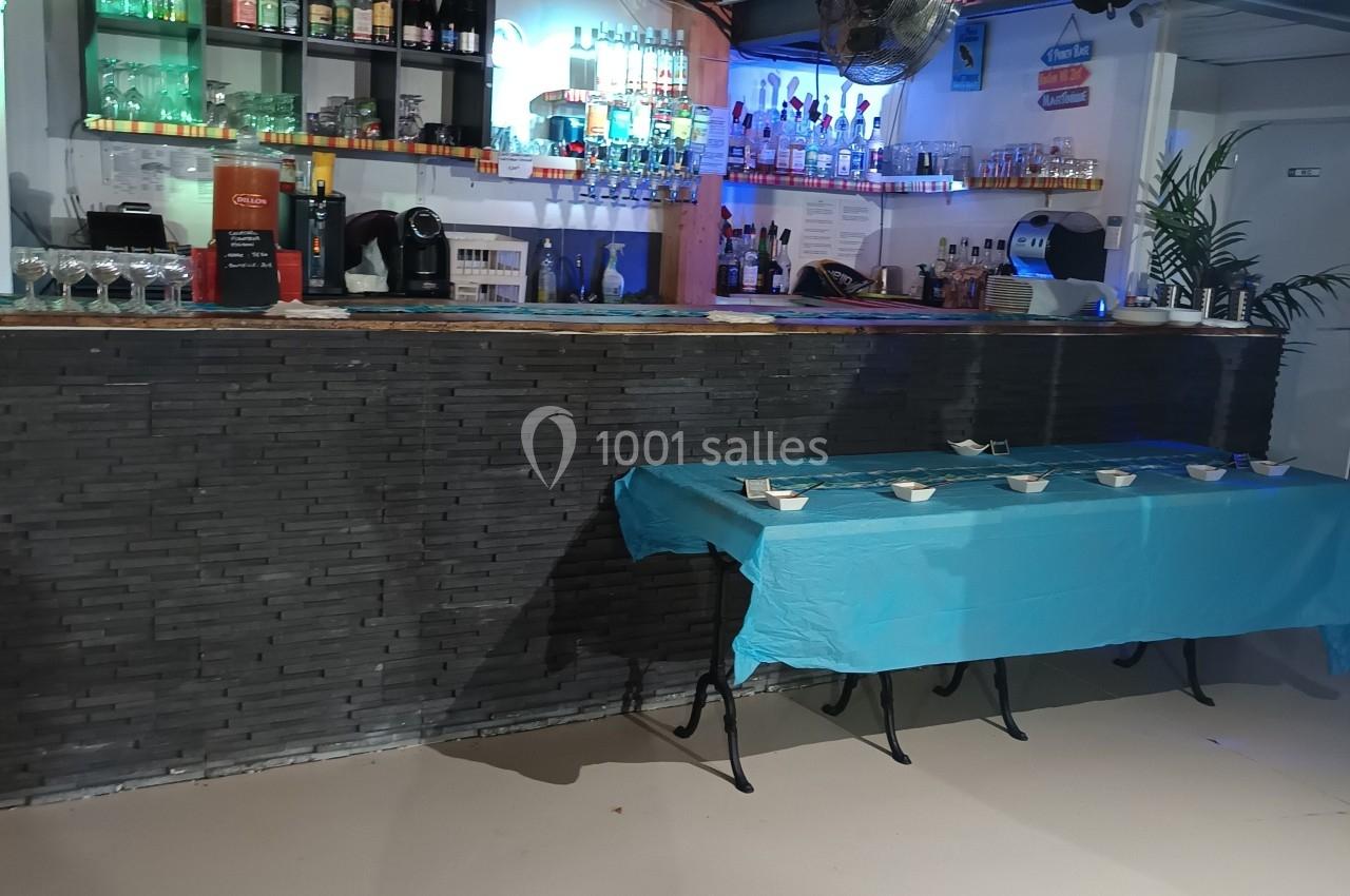 Bar avec comptoir en pierre, étagères de bouteilles éclairées, table avec nappes bleues et vaisselle disposée.