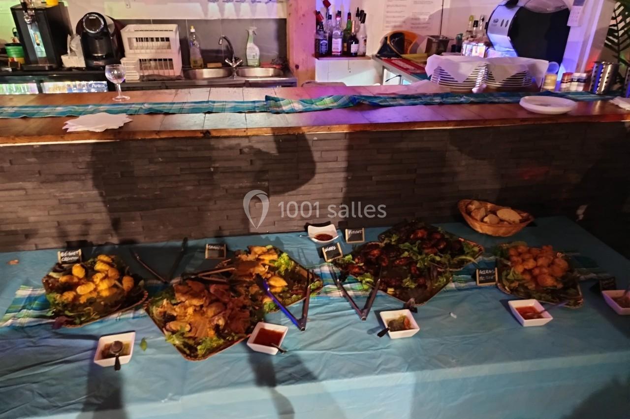 Buffet avec divers plats et accompagnements disposés sur une table recouverte d'une nappe bleue, bar en arrière-plan.