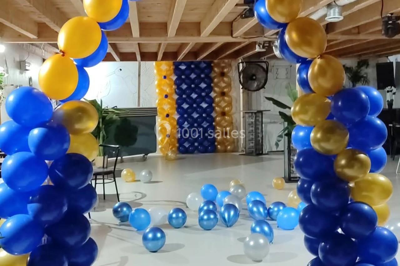 Décoration de salle avec des arches de ballons bleus et jaunes, et des ballons dispersés au sol.