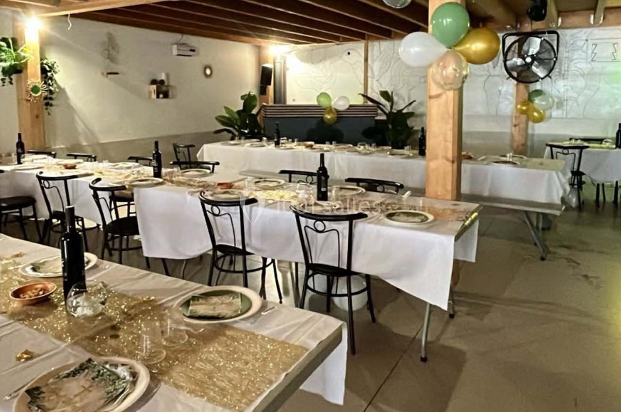 Salle décorée pour un événement, avec tables dressées, nappes blanches, ballons et plantes en arrière-plan.