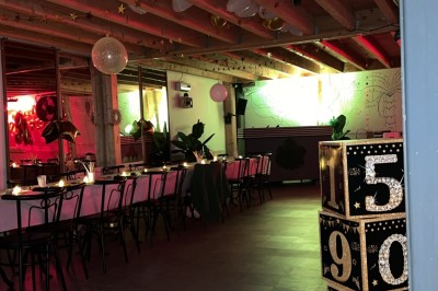 Salle décorée pour une fête avec une longue table dressée, bougies, confettis et ballons dorés.