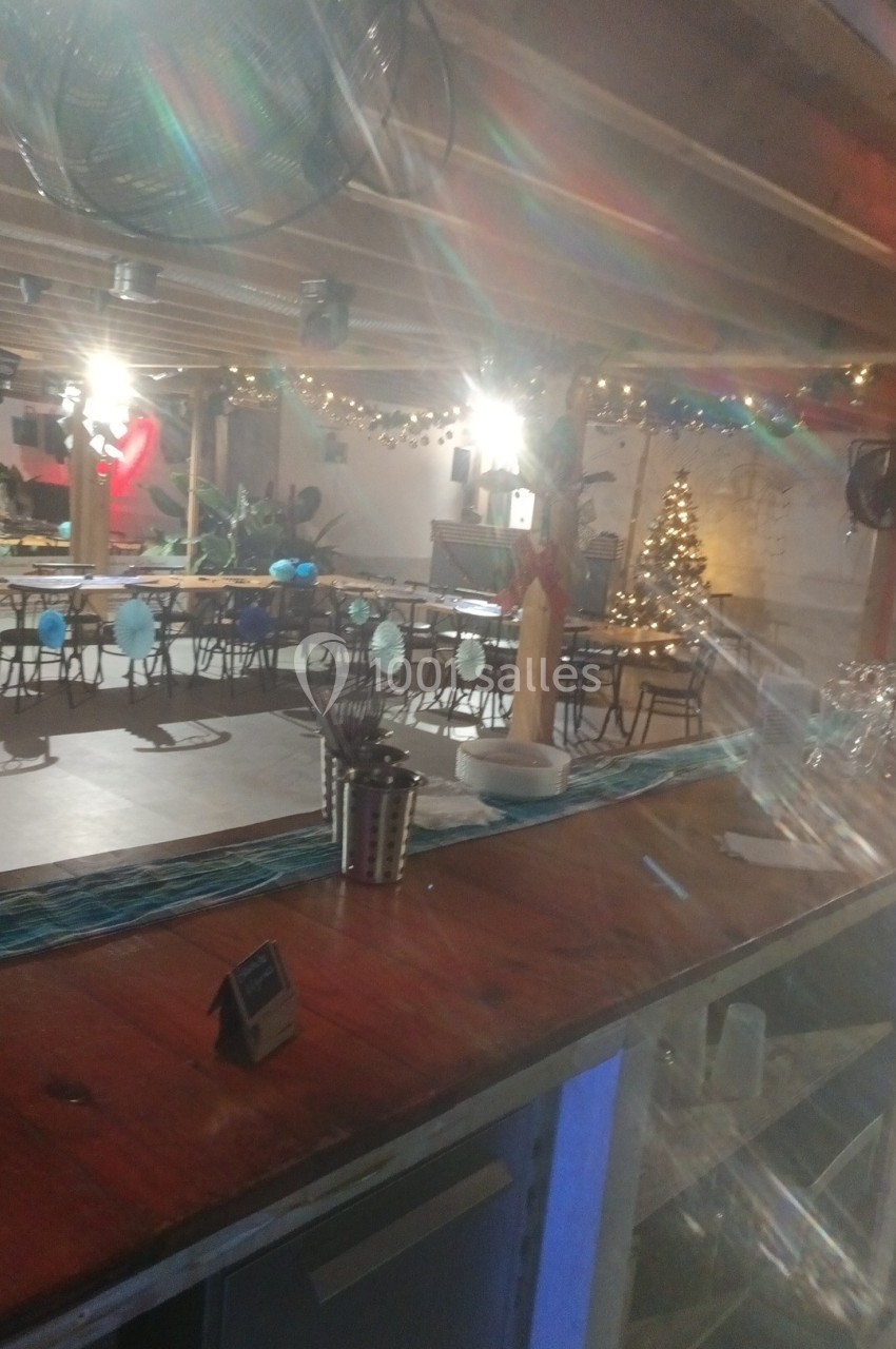 Salle décorée pour une fête avec guirlandes lumineuses, sapin de Noël et tables alignées sous un plafond en bois.