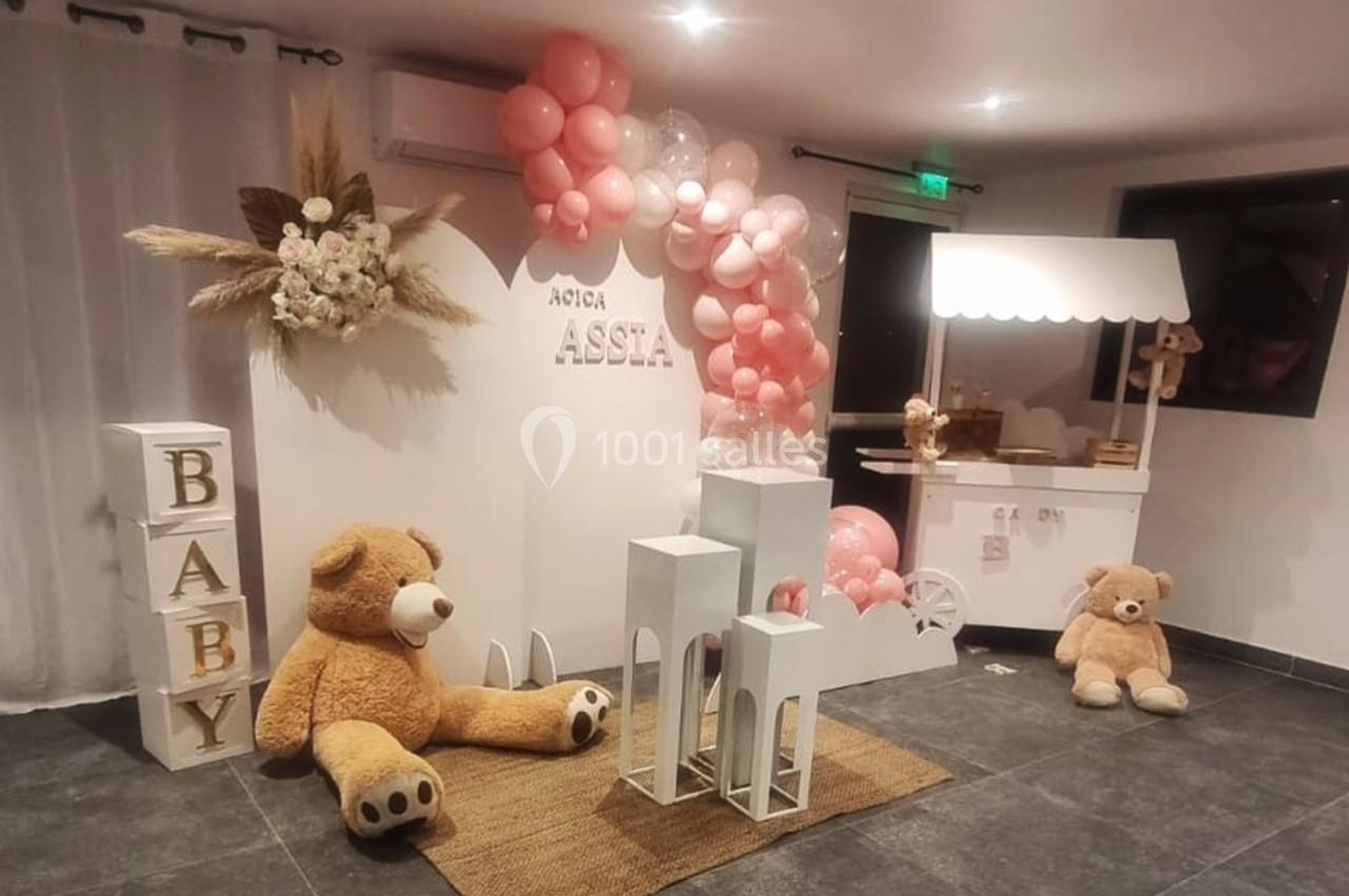 Décoration de fête avec ballons roses, oursons en peluche, cubes ’BABY’ et stand blanc dans une pièce lumineuse.