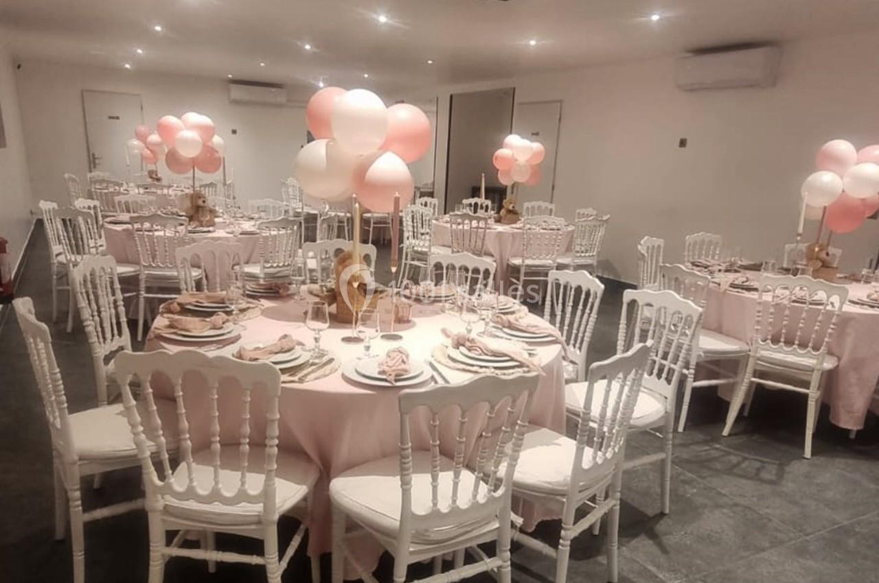Salle décorée pour un événement avec des tables rondes, nappes roses, chaises blanches et ballons roses et blancs.