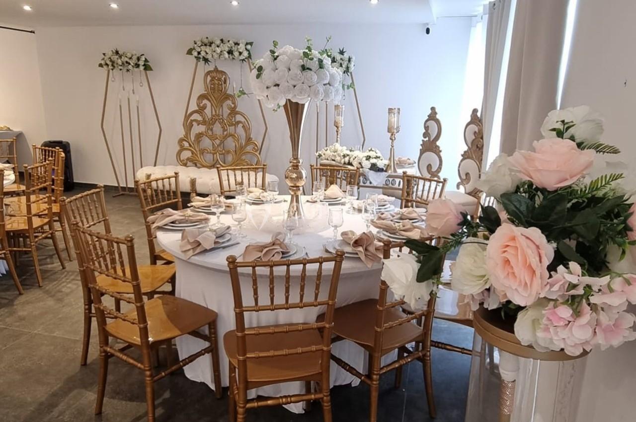 Salle de réception décorée avec des tables rondes, des chaises dorées et des arrangements floraux blancs et roses.