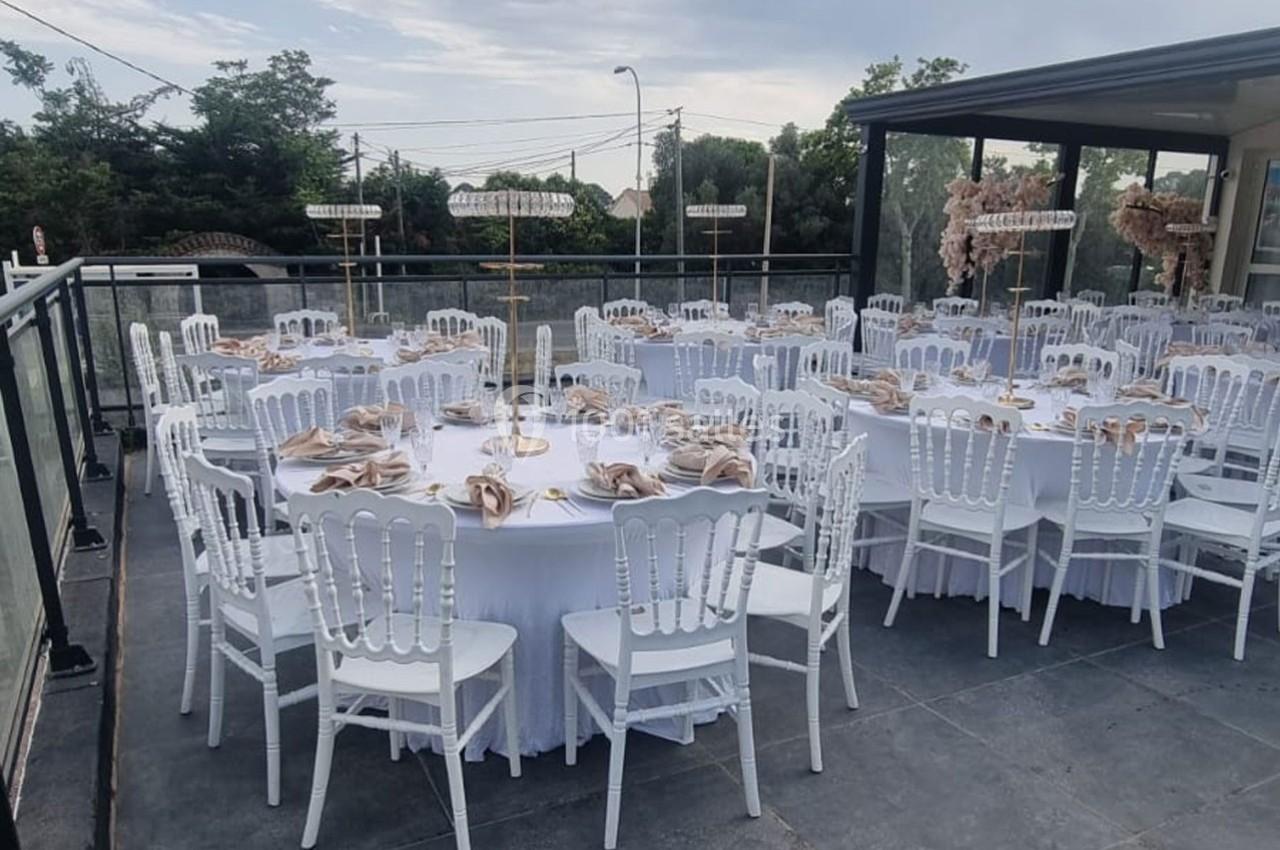 Tables rondes dressées avec nappes blanches et chaises blanches sur une terrasse extérieure, décorées pour un événement.