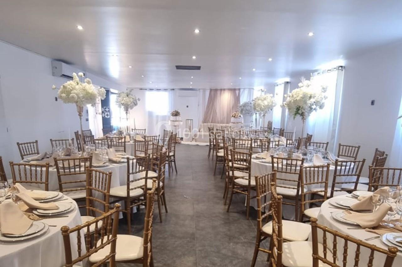 Salle de réception décorée avec des tables rondes, nappes blanches, chaises dorées et centres de table fleuris.