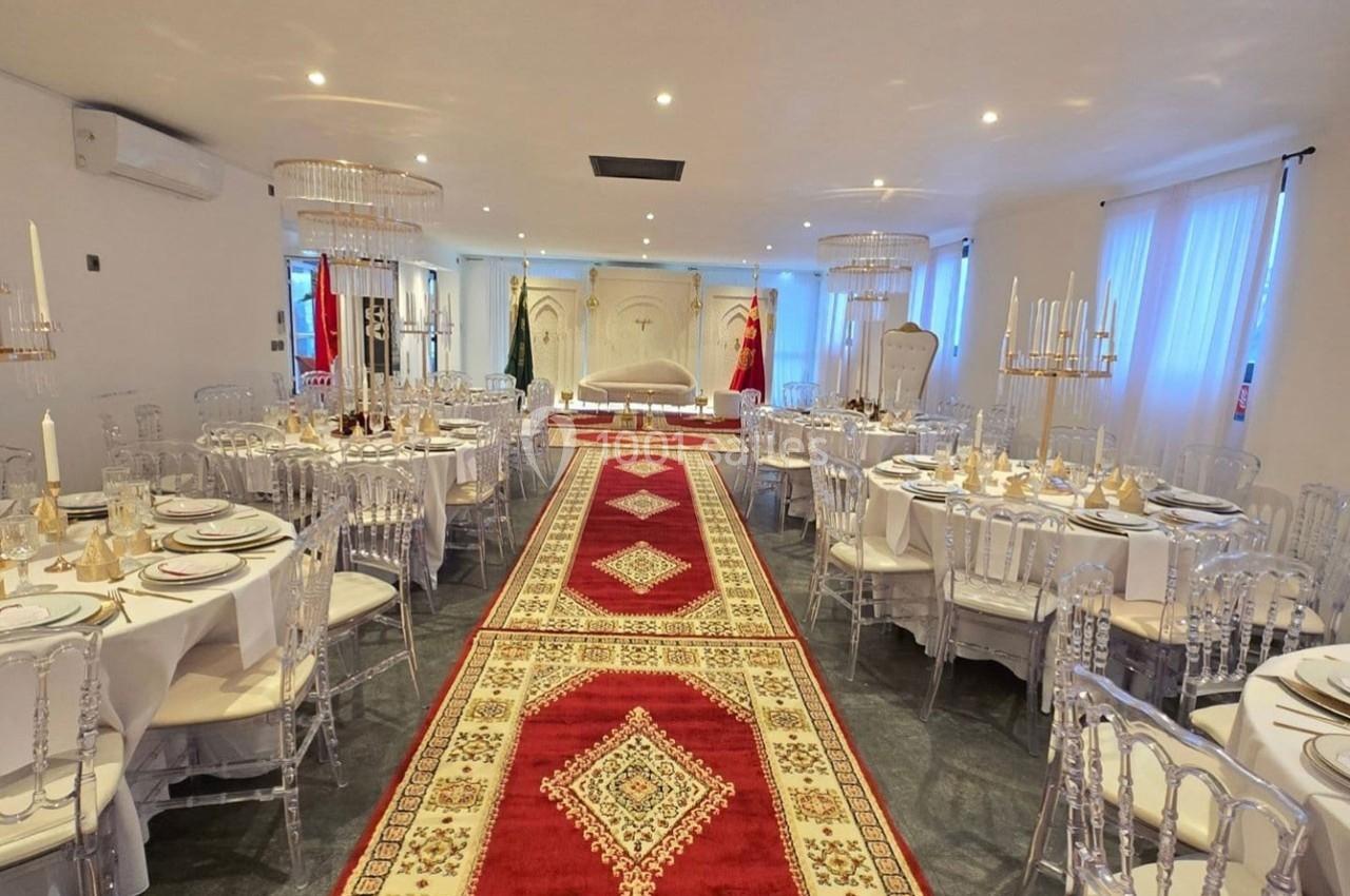 Salle de réception décorée avec des tables rondes dressées, un tapis rouge central et un espace scénique au fond.