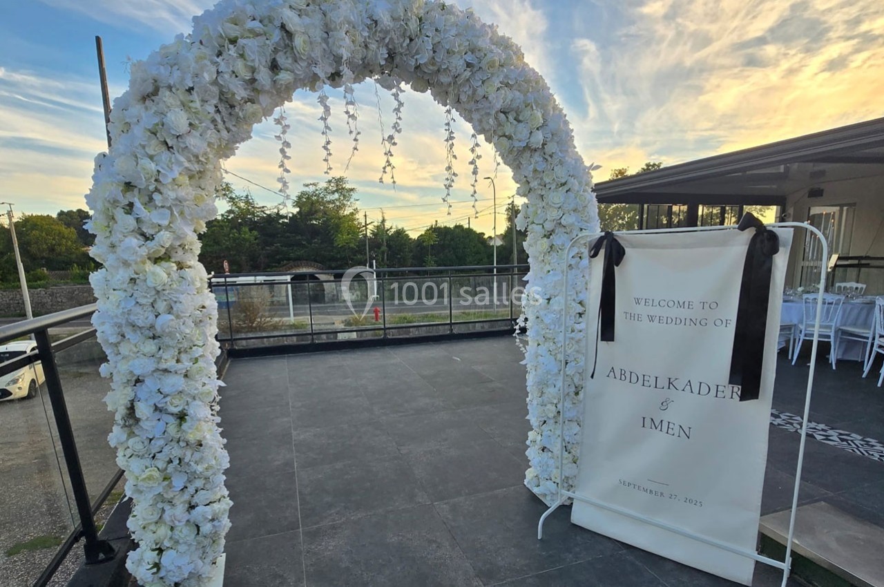 Arc fleuri blanc sur une terrasse extérieure au coucher du soleil, avec un panneau de bienvenue pour un mariage.